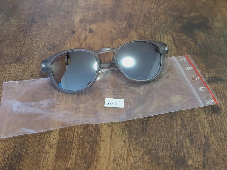 May include: Gray round sunglasses with dark lenses rest on a clear plastic bag. The sunglasses have a matte finish and a keyhole bridge. A small white tag with the number "105" is visible.