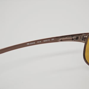 May include: Close-up of brown sunglasses with yellow lenses. The frame has the markings "RB 3343 014 60012 3N". The sunglasses are set against a white background.