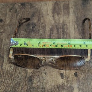 Peut inclure: Lunettes de soleil &agrave; monture dor&eacute;e avec verres bruns, d'environ 14 cm de large. Le logo Ray-Ban est visible sur le verre. Les branches ont un motif. Les lunettes sont pos&eacute;es sur une surface en bois.