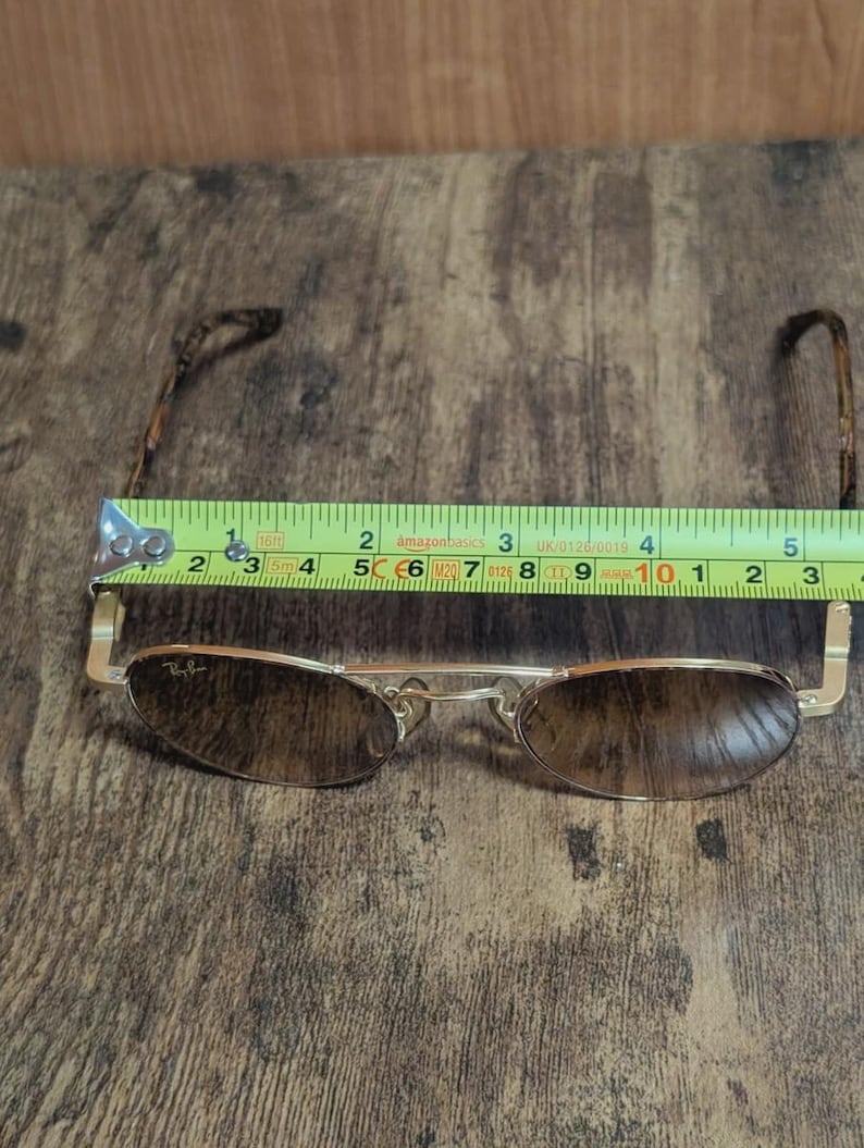 Peut inclure: Lunettes de soleil &agrave; monture dor&eacute;e avec verres bruns, d'environ 14 cm de large. Le logo Ray-Ban est visible sur le verre. Les branches ont un motif. Les lunettes sont pos&eacute;es sur une surface en bois.
