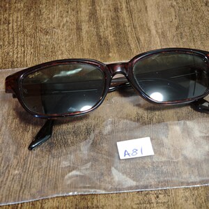 May include: Dark brown tortoiseshell sunglasses with dark lenses. The sunglasses have a rectangular frame and black arms. The Ray-Ban logo is visible on the upper left lens. A small white tag with the text "A81" is in the lower right corner.