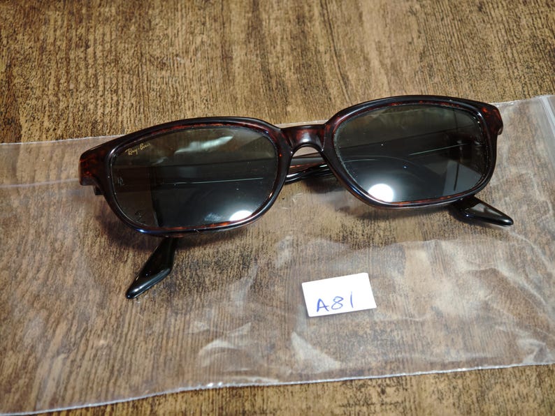 May include: Dark brown tortoiseshell sunglasses with dark lenses. The sunglasses have a rectangular frame and black arms. The Ray-Ban logo is visible on the upper left lens. A small white tag with the text "A81" is in the lower right corner.
