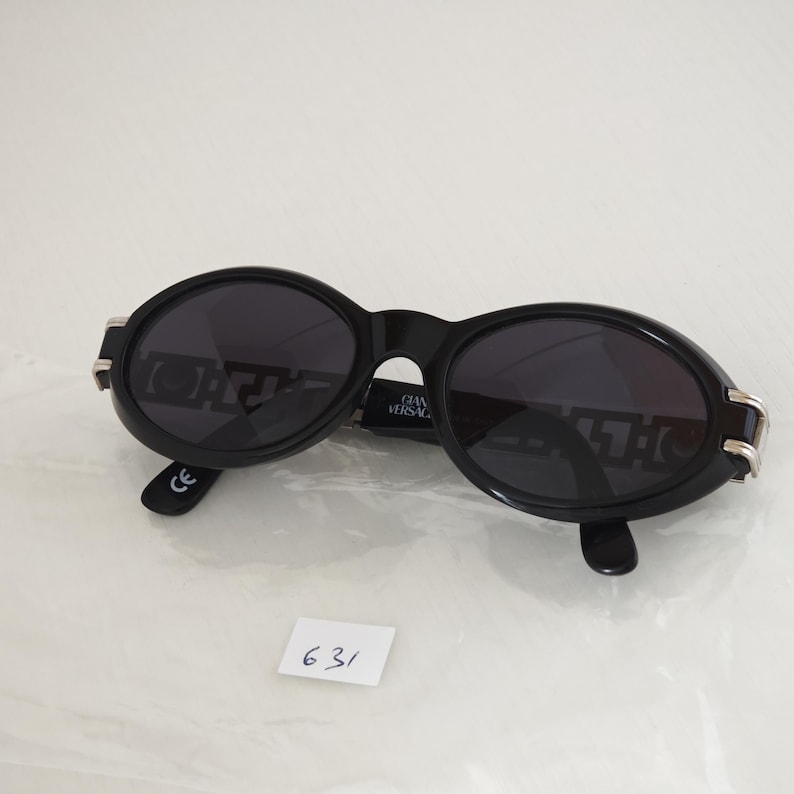 Puede incluir: Gafas de sol ovaladas negras con lentes oscuras y detalles met&aacute;licos plateados en las bisagras exteriores. La patilla interior muestra el texto GIANNI VERSACE. La montura tiene un acabado liso y brillante con una marca CE en la punta.