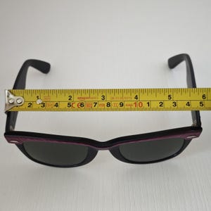 Puede incluir: Gafas de sol negras con lentes oscuras y un acento morado a lo largo del marco superior. Una cinta m&eacute;trica amarilla est&aacute; colocada sobre el frente, mostrando un ancho de aproximadamente 14 cm desde el borde exterior de una bisagra a la otra.