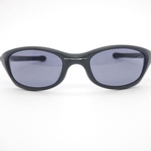 May include: Black sunglasses with gray lenses. The sunglasses have a curved, oval shape and the word "OAKLEY" is printed in white on the bridge. The frames are matte black.