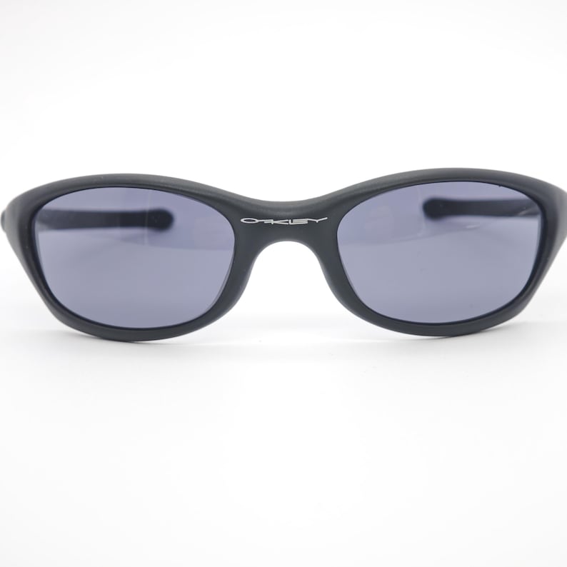 May include: Black sunglasses with gray lenses. The sunglasses have a curved, oval shape and the word "OAKLEY" is printed in white on the bridge. The frames are matte black.