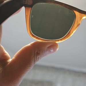 May include: Close-up of sunglasses with a tortoiseshell frame and dark green lenses. The sunglasses are held by a hand, with the focus on the frame's color and the lenses' shape. The lenses have a slight scratch.