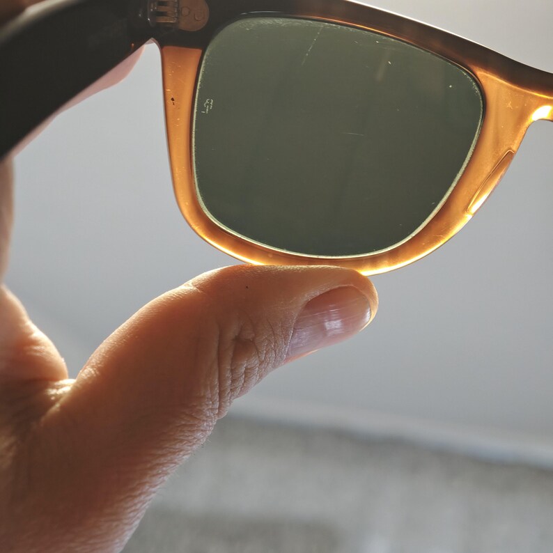 May include: Close-up of sunglasses with a tortoiseshell frame and dark green lenses. The sunglasses are held by a hand, with the focus on the frame's color and the lenses' shape. The lenses have a slight scratch.