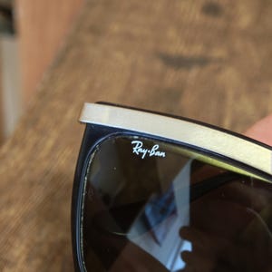 Peut inclure: Lunettes de soleil Ray-Ban noires avec une barre sup&eacute;rieure argent&eacute;e. Le logo Ray-Ban est visible sur le verre. Les lunettes ont une monture rectangulaire et des verres fonc&eacute;s.