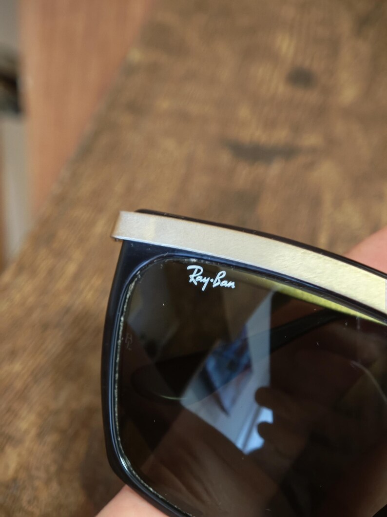 Peut inclure: Lunettes de soleil Ray-Ban noires avec une barre sup&eacute;rieure argent&eacute;e. Le logo Ray-Ban est visible sur le verre. Les lunettes ont une monture rectangulaire et des verres fonc&eacute;s.