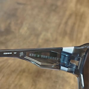 May include: Close-up of a pair of black sunglasses with the text "OFFSHOOT" visible on the temple. The frame is a translucent black with silver accents. The model number "OO9190-05 127" is also visible.