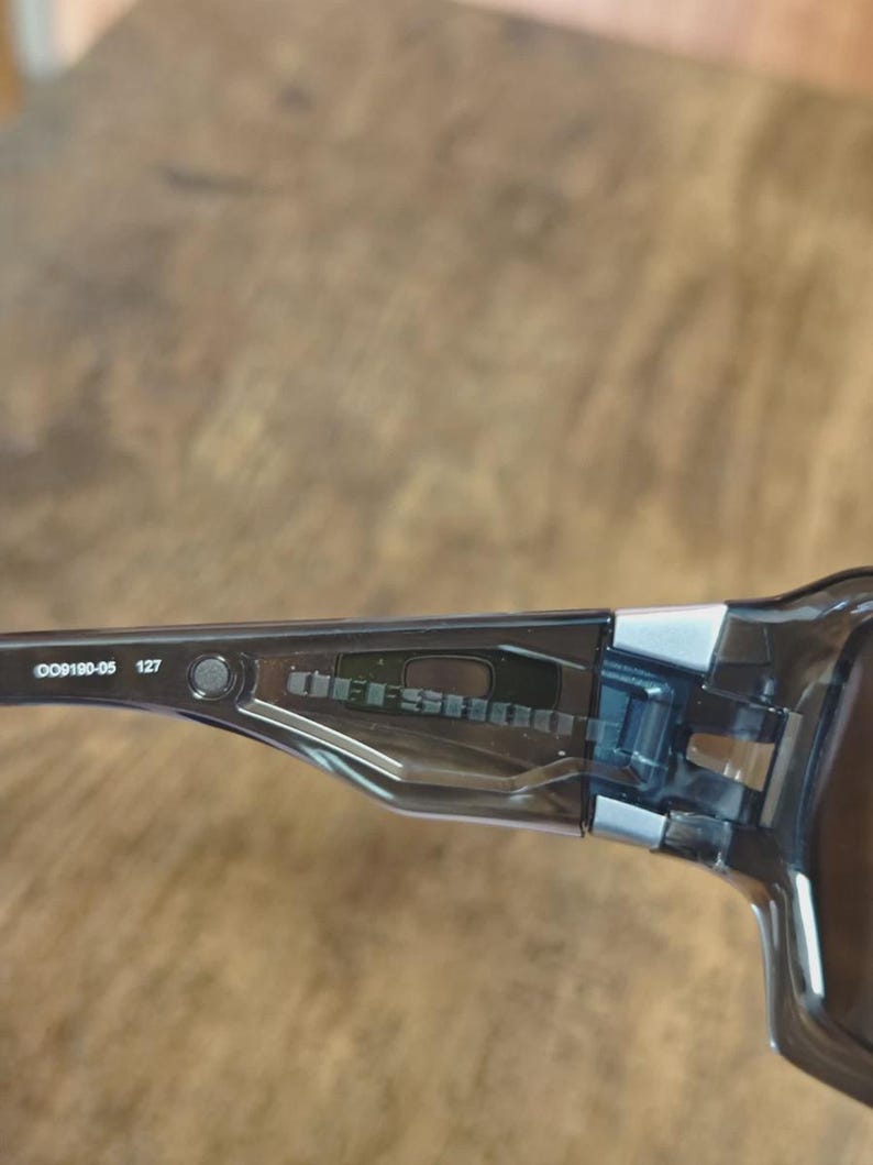May include: Close-up of a pair of black sunglasses with the text "OFFSHOOT" visible on the temple. The frame is a translucent black with silver accents. The model number "OO9190-05 127" is also visible.