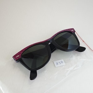 Puede incluir: Gafas de sol con lentes oscuras y montura negra con una barra superior texturizada en color morado. La montura incluye peque&ntilde;os detalles plateados en las esquinas. Se incluye una peque&ntilde;a etiqueta blanca con el texto escrito a mano K37.