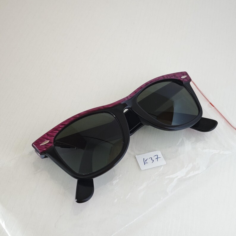 Puede incluir: Gafas de sol con lentes oscuras y montura negra con una barra superior texturizada en color morado. La montura incluye peque&ntilde;os detalles plateados en las esquinas. Se incluye una peque&ntilde;a etiqueta blanca con el texto escrito a mano K37.