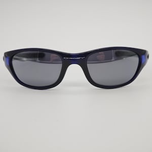 May include: Dark blue Oakley sunglasses with grey lenses. The sunglasses have a curved frame and the Oakley logo is visible on the bridge. The temples have a blue accent.