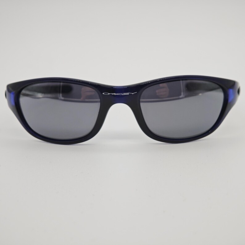 May include: Dark blue Oakley sunglasses with grey lenses. The sunglasses have a curved frame and the Oakley logo is visible on the bridge. The temples have a blue accent.