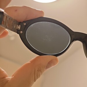Puede incluir: Gafas de sol ovaladas de color negro con lentes oscuras y un mecanismo de bisagra met&aacute;lico con tornillos visibles que conectan la montura con la patilla. La montura tiene un acabado liso y brillante y una silueta redondeada y cl&aacute;sica.