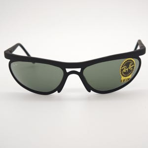 May include: Black sunglasses with green lenses. The sunglasses have a curved frame and a small logo on the lens. The Ray-Ban logo is visible on the right lens. The sunglasses are on a white background.