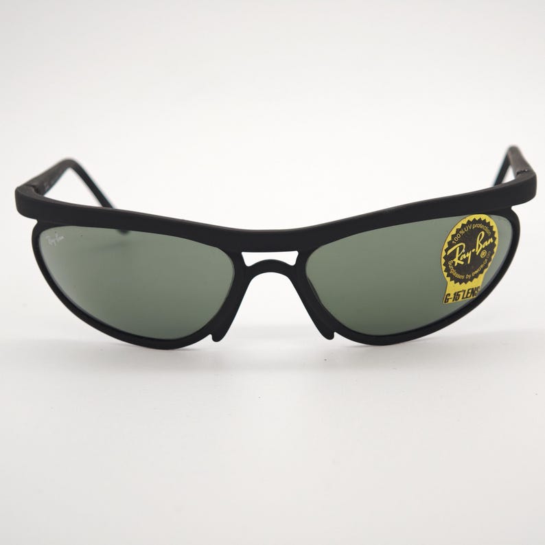 May include: Black sunglasses with green lenses. The sunglasses have a curved frame and a small logo on the lens. The Ray-Ban logo is visible on the right lens. The sunglasses are on a white background.
