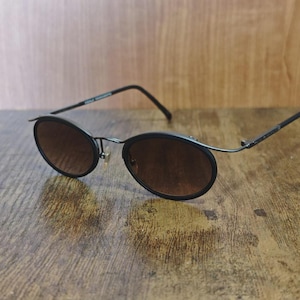 May include: Black oval sunglasses with brown tinted lenses. The sunglasses have a thin, dark metal frame and are resting on a wooden surface. The arms of the sunglasses curve down at the ends.