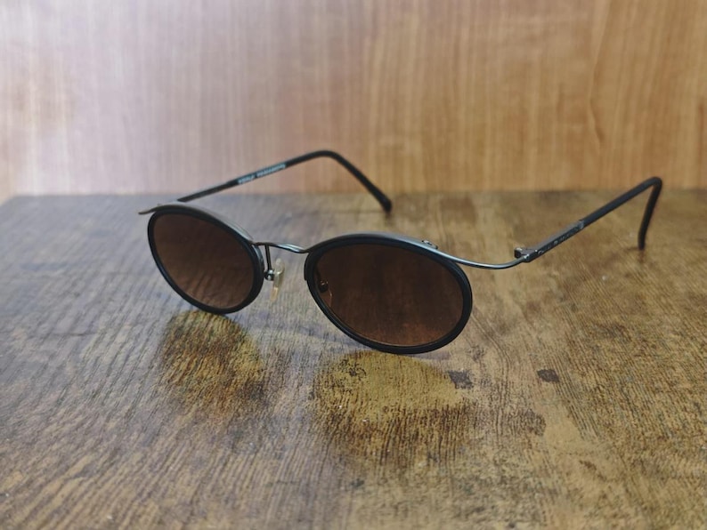 May include: Black oval sunglasses with brown tinted lenses. The sunglasses have a thin, dark metal frame and are resting on a wooden surface. The arms of the sunglasses curve down at the ends.