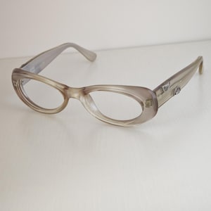 May include: Translucent taupe-colored eyeglasses with an oval frame shape. The temples feature small, silver-toned metallic accents embedded near the hinges. The frames have a smooth, polished finish and clear lenses, showcasing a simple design.