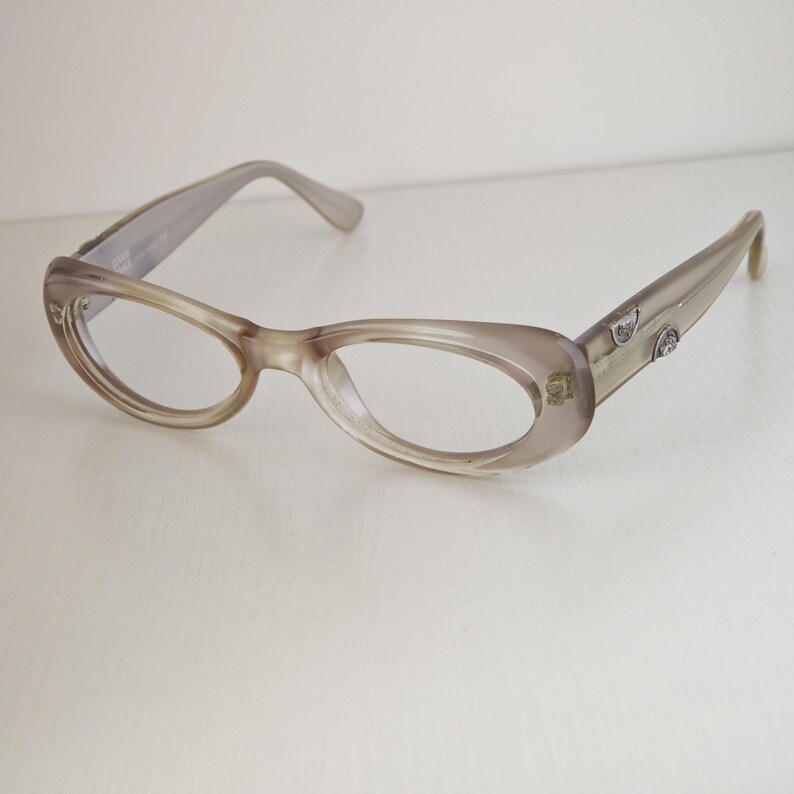 May include: Translucent taupe-colored eyeglasses with an oval frame shape. The temples feature small, silver-toned metallic accents embedded near the hinges. The frames have a smooth, polished finish and clear lenses, showcasing a simple design.