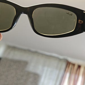 Może przedstawiać: Czarne okulary przeciwsłoneczne z prostokątną oprawką i zielonymi szkłami. Okulary przeciwsłoneczne mają biały akcent na g&oacute;rze oprawki. Logo Ray-Ban jest widoczne na prawej soczewce.