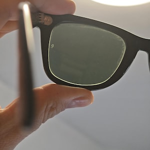 May include: Black sunglasses with dark green lenses. The sunglasses have a classic rectangular frame design. The image shows a close-up view of the sunglasses, with a focus on the lenses and frame.