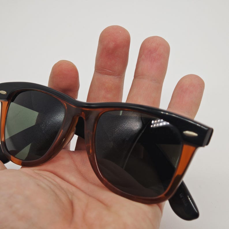 May include: Sunglasses with a classic design. The frames are black on top and transition to a brown tortoise shell pattern. The lenses are dark, and the sunglasses are held in a hand.
