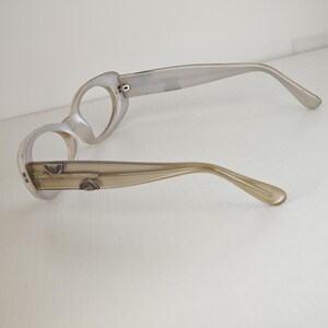 May include: A pair of translucent beige eyeglasses with round frames and thick temples. The temples feature small, metallic, semi-circular decorative accents embedded into the side. The frames are empty, lacking lenses, and have a smooth, glossy finish.