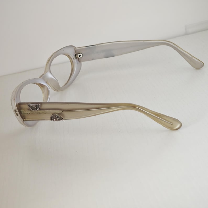 May include: A pair of translucent beige eyeglasses with round frames and thick temples. The temples feature small, metallic, semi-circular decorative accents embedded into the side. The frames are empty, lacking lenses, and have a smooth, glossy finish.