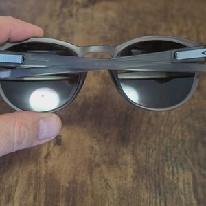 May include: Gray sunglasses with dark lenses are held up, revealing the inner frame and hinges. The arms have a light blue accent with a white logo. The sunglasses are set against a wooden background.