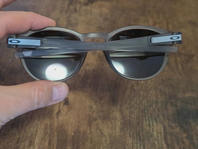 May include: Gray sunglasses with dark lenses are held up, revealing the inner frame and hinges. The arms have a light blue accent with a white logo. The sunglasses are set against a wooden background.