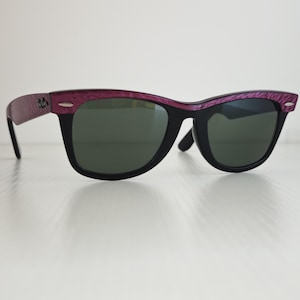 Puede incluir: Gafas de sol con montura negra y lentes de color verde oscuro. La barra superior y las patillas presentan un acabado texturizado en color morado intenso. Peque&ntilde;os detalles met&aacute;licos plateados son visibles en las esquinas frontales de la montura, y el logotipo de Ray-Ban est&aacute; impreso en la patilla.