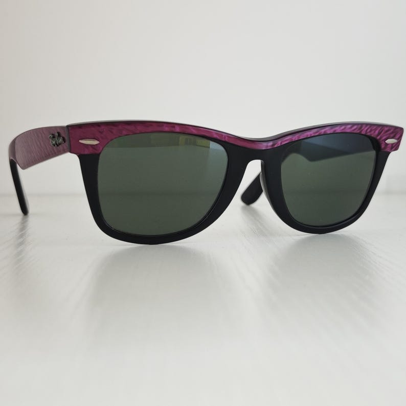 Puede incluir: Gafas de sol con montura negra y lentes de color verde oscuro. La barra superior y las patillas presentan un acabado texturizado en color morado intenso. Peque&ntilde;os detalles met&aacute;licos plateados son visibles en las esquinas frontales de la montura, y el logotipo de Ray-Ban est&aacute; impreso en la patilla.
