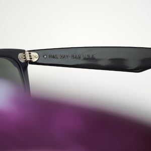 Puede incluir: Gafas de sol negras con una bisagra met&aacute;lica y el texto B&L RAY-BAN U.S.A. grabado en la patilla interior. La montura est&aacute; fabricada con un material oscuro y liso con un mecanismo de bisagra visible que conecta la patilla con el marco.