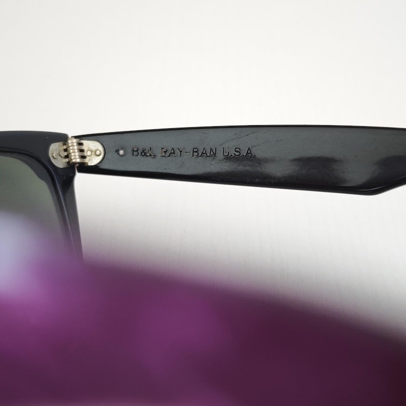Puede incluir: Gafas de sol negras con una bisagra met&aacute;lica y el texto B&L RAY-BAN U.S.A. grabado en la patilla interior. La montura est&aacute; fabricada con un material oscuro y liso con un mecanismo de bisagra visible que conecta la patilla con el marco.