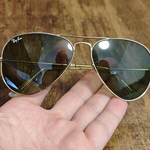 May include: Gold-framed aviator sunglasses with dark lenses. The sunglasses have a double bridge and the Ray-Ban logo is visible on the upper left lens. The lenses reflect the surrounding environment. The sunglasses are resting on a hand.