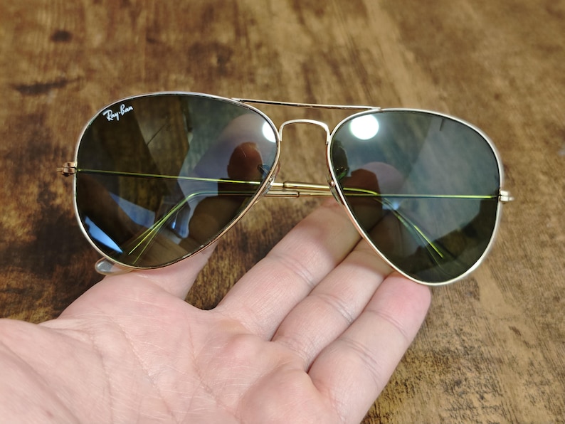 May include: Gold-framed aviator sunglasses with dark lenses. The sunglasses have a double bridge and the Ray-Ban logo is visible on the upper left lens. The lenses reflect the surrounding environment. The sunglasses are resting on a hand.