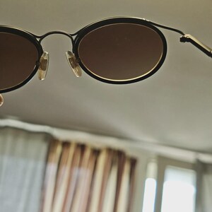 May include: Black oval-framed sunglasses with brown lenses. The sunglasses have a thin, black metal frame and a classic design. The lenses are a warm brown color, and the nose pads are clear.