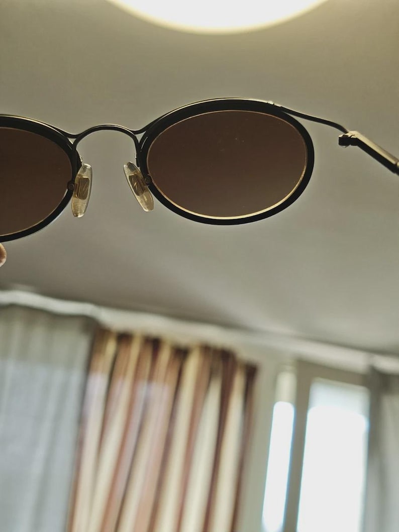 May include: Black oval-framed sunglasses with brown lenses. The sunglasses have a thin, black metal frame and a classic design. The lenses are a warm brown color, and the nose pads are clear.