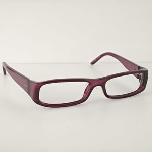 May include: A pair of rectangular spectacles with a translucent deep purple plastic frame. The temples feature the Gucci brand name embossed in small silver lettering on the side. The glasses have a smooth, polished finish and clear lenses.