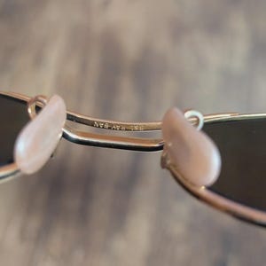 May include: Close-up of gold-framed sunglasses with dark lenses. The nose pads are a light pink color. The text "RB RAY-BAN" is visible on the frame.