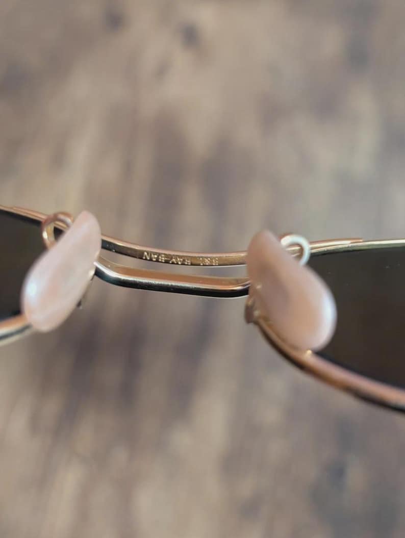 May include: Close-up of gold-framed sunglasses with dark lenses. The nose pads are a light pink color. The text "RB RAY-BAN" is visible on the frame.