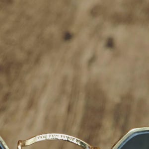 May include: Close-up of gold-rimmed sunglasses with dark lenses. The bridge of the glasses has the text "BALMAIN PARIS" etched into the metal. The background is a blurred, textured brown.
