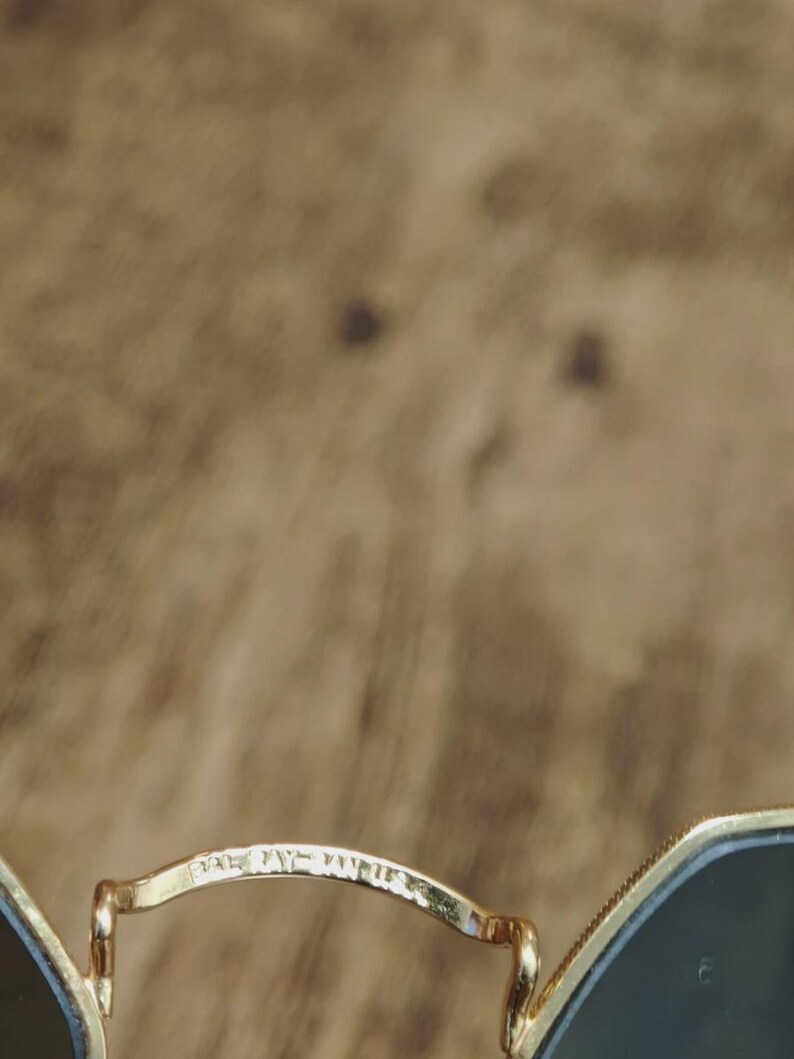 May include: Close-up of gold-rimmed sunglasses with dark lenses. The bridge of the glasses has the text "BALMAIN PARIS" etched into the metal. The background is a blurred, textured brown.