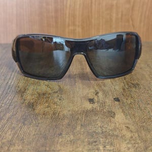 May include: Black sunglasses with dark tinted lenses. The sunglasses have a curved, wraparound design and are set against a wooden surface. The lenses reflect the surrounding environment.