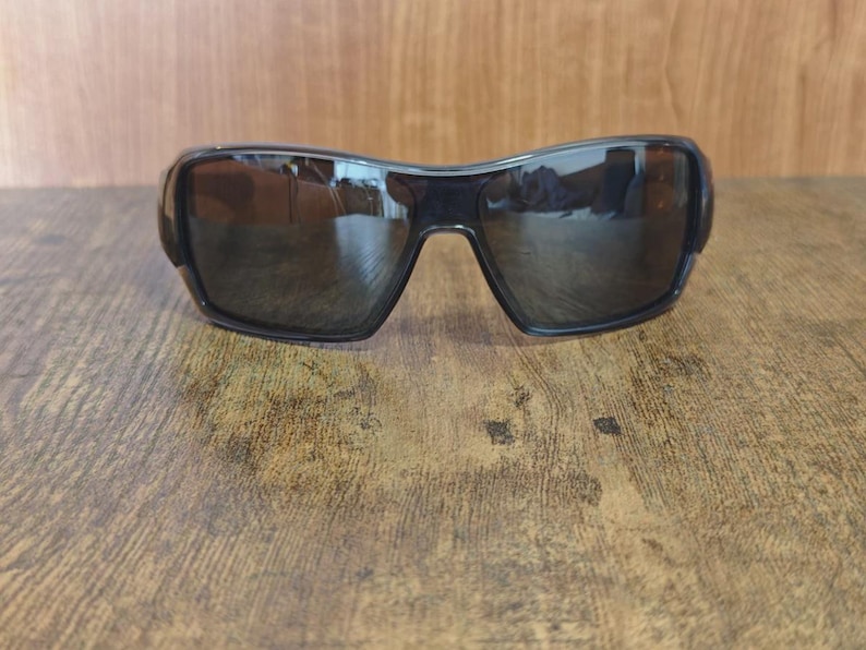 May include: Black sunglasses with dark tinted lenses. The sunglasses have a curved, wraparound design and are set against a wooden surface. The lenses reflect the surrounding environment.