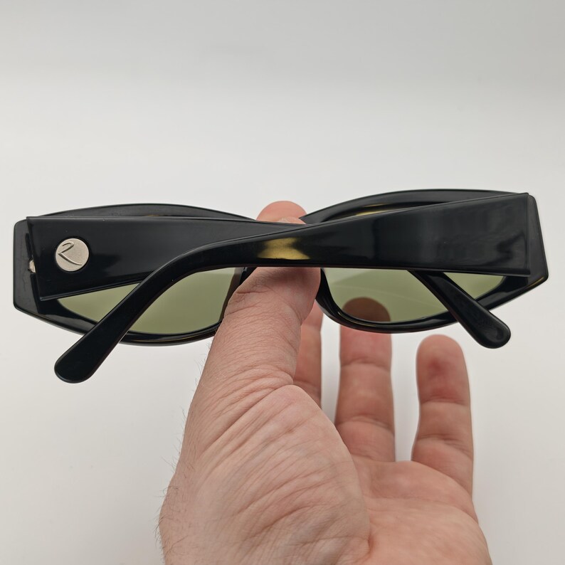 Może przedstawiać: Czarne okulary przeciwsłoneczne o geometrycznym designie. Okulary mają grubą, prostokątną oprawkę ze srebrnym akcentem z boku. Soczewki są jasnozielone.